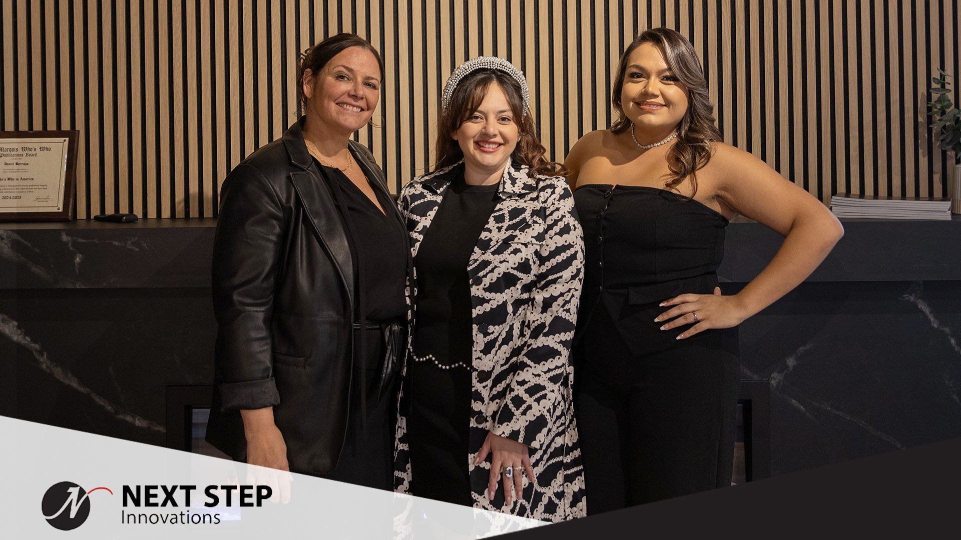 Image of three female members of the Next Step Innovations team standing in front of a slat wood fireplace backdrop