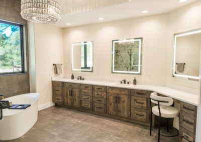A luxury master bathroom featuring a long wood double vanity with white quartz countertops, two backlit LED mirrors, a freestanding soaking tub, and a decorative circular chandelier.